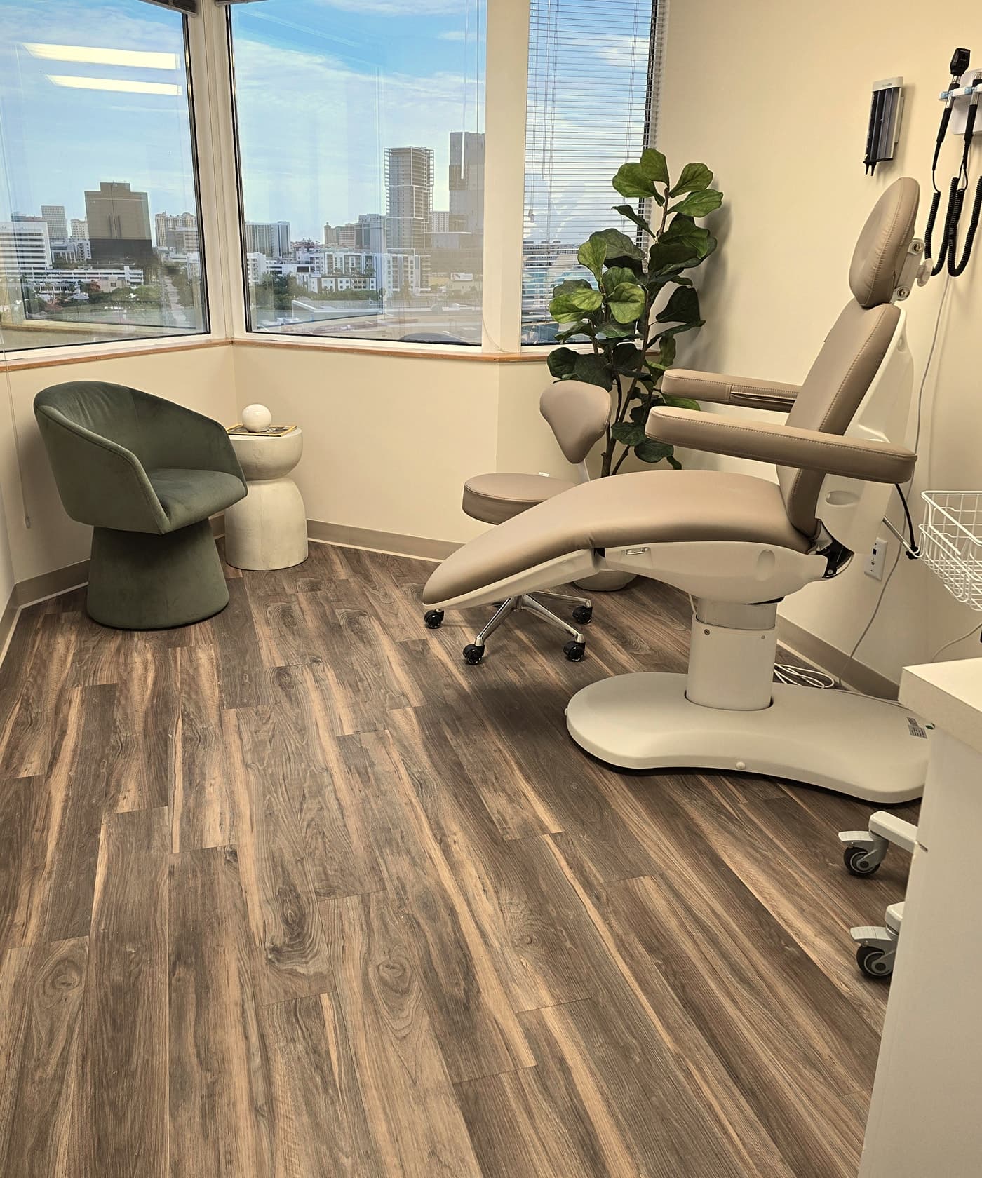 A GreenTree exam room with a tan exam chair, green accent chair, fiddle-leaf fig, and corner windows overlooking the West Palm Beach skyline